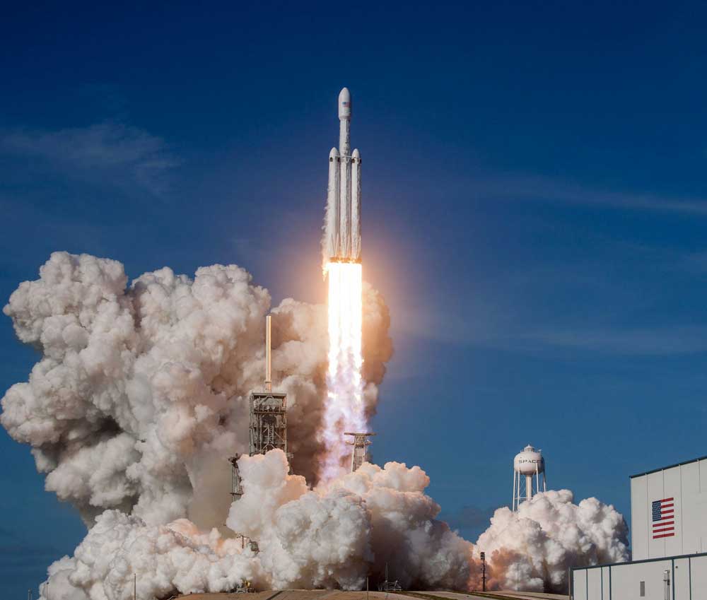 McCollisters A rocket launches into the sky, surrounded by billowing smoke and flames, leaving its launch pad. The sky is clear and blue, and a building with a U.S. flag is visible in the foreground.
