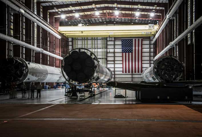 McCollisters Three large rocket boosters are positioned horizontally inside a spacious industrial facility, highlighting advanced logistics in the aviation sector, with an American flag on the back wall and several workers standing nearby.