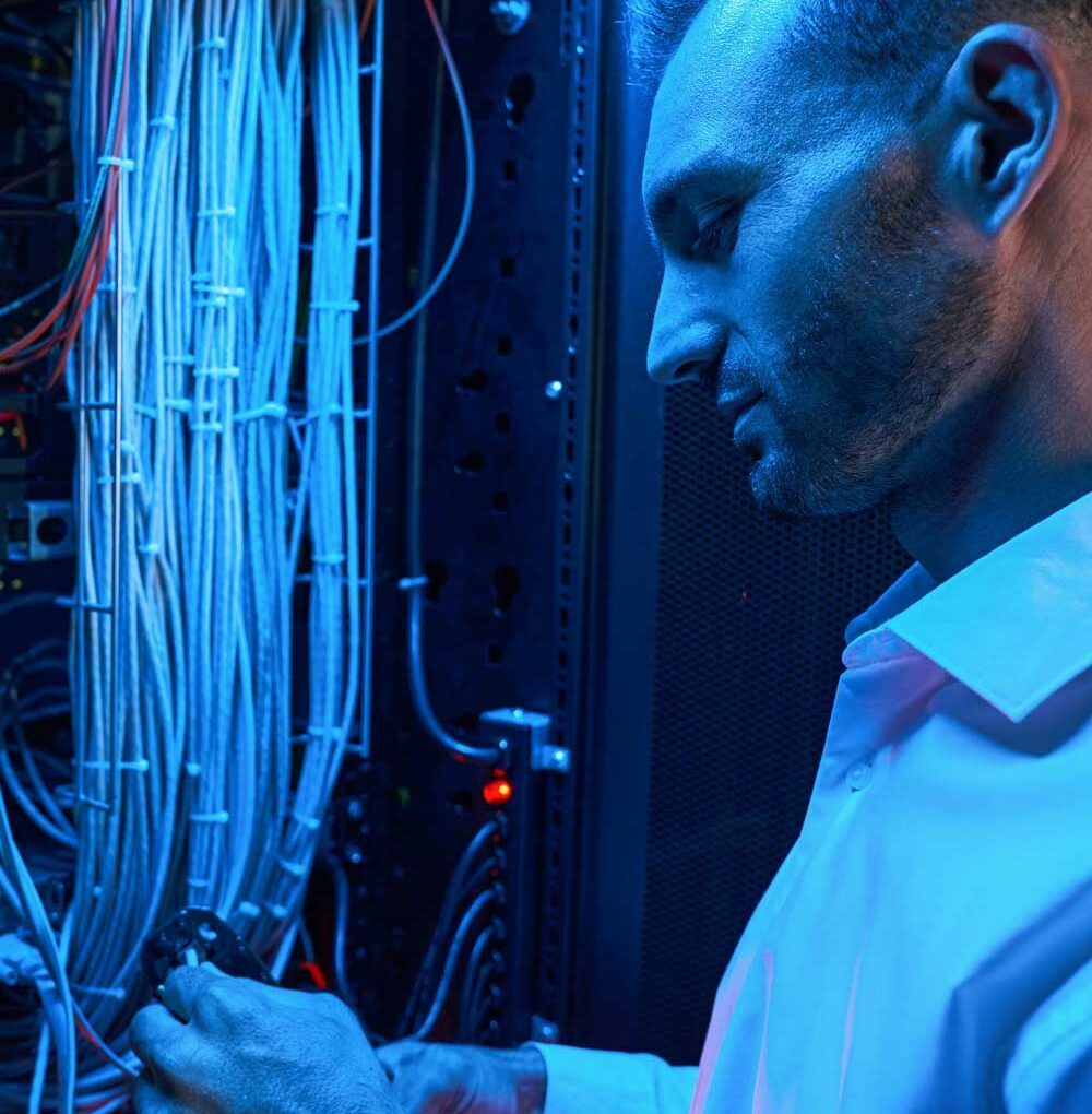 McCollisters A man in a white shirt works on a server rack filled with numerous cables, illuminated by blue and red lights, providing Technical Services in a data center or server room.