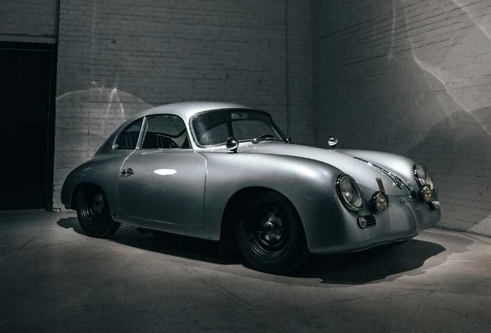McCollisters A classic silver vintage auto with rounded edges is parked in a dimly lit room with white brick walls and concrete floors. The car, often admired by logistics enthusiasts, features prominent round headlights and a smooth, curved body design.