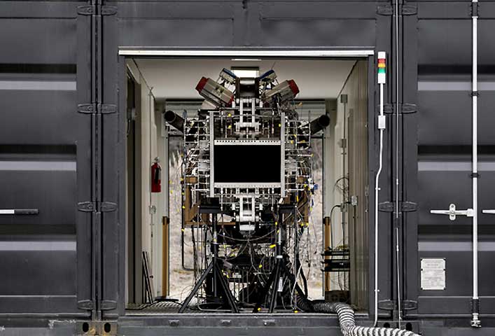 McCollisters A complex scientific instrument with numerous wires and metal parts, used in aviation research, is housed inside an open shipping container—highlighting advanced logistics. A black monitor sits at the center of the device, visible through a doorway.