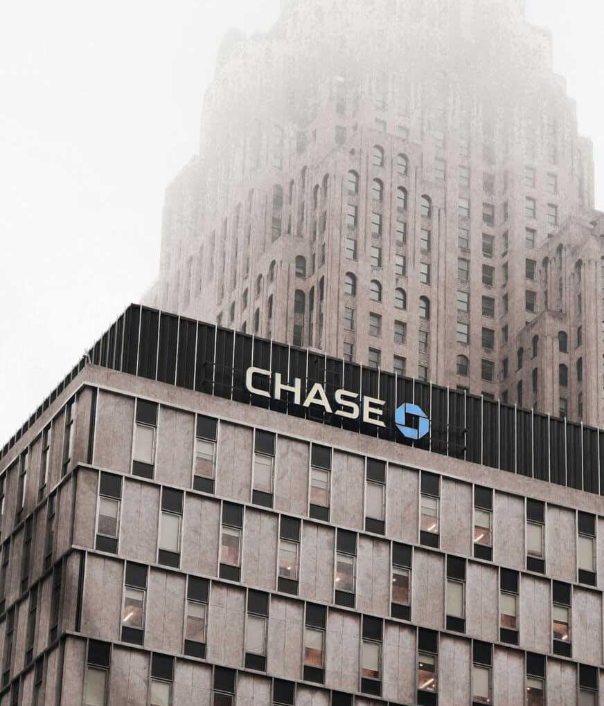 McCollisters A multistory building with the Chase bank logo on its facade, symbolizing the world of finance and banking, stands before a taller skyscraper partially obscured by fog in this overcast, urban scene.