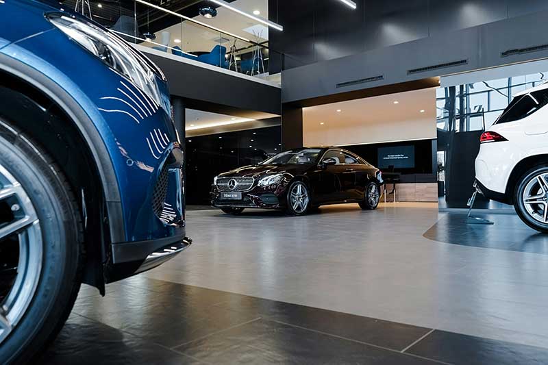 A modern car showroom with a black luxury sedan displayed in the center, surrounded by other vehicles. The space features sleek design, large windows, and a mezzanine level above.