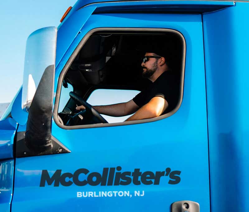 McCollisters A man wearing sunglasses drives a blue truck with McCollisters Burlington, NJ written on the door. The photo is taken from outside the drivers side window on a sunny day.