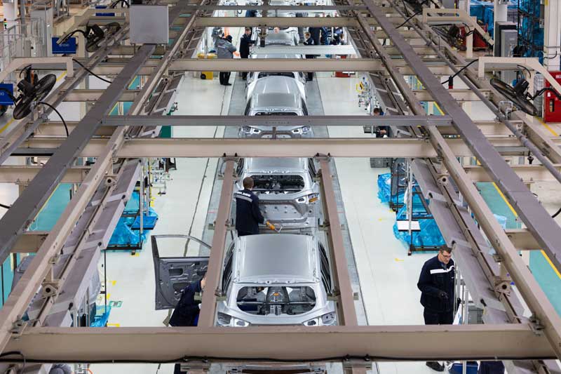 Overhead view of an automotive assembly line with workers assembling silver car bodies, surrounded by blue machinery and structural beams inside a brightly lit factory.