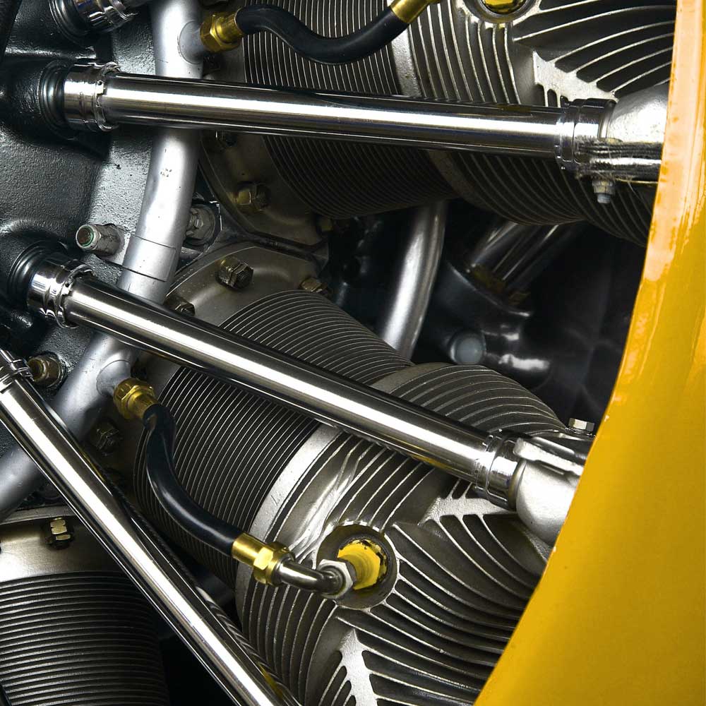 McCollisters Close-up of a vintage radial aircraft engine showing metal cylinders, connecting rods, and black hoses, with part of a yellow engine cowling visible on the right side.