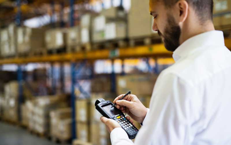 McCollisters A person in a white shirt uses a handheld electronic device with a stylus in a warehouse filled with shelves of stacked boxes and packages.