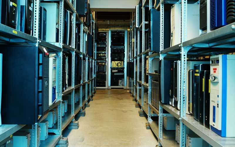 McCollisters Rows of metal shelves holding computer servers and hardware components line a hallway in a data center or server room, with cables and equipment visible in the background.