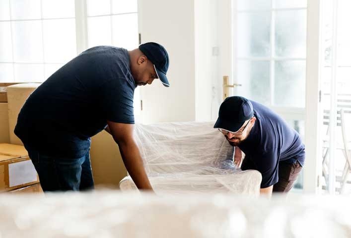 McCollisters Two men wearing dark shirts and caps are lifting a plastic-wrapped armchair in a bright residential space with moving boxes, large windows, and ongoing transportation activities in the background.