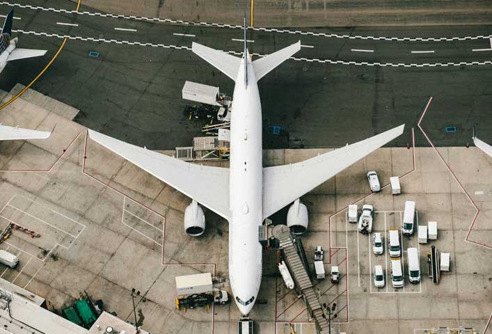 McCollisters Aerial view of a large white airplane parked at an airport gate, with transportation vehicles and equipment positioned nearby on the tarmac. The wings and engines are clearly visible.