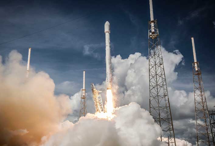 McCollisters A rocket launches into the sky, surrounded by clouds of smoke and supported by metal towers, against a backdrop of dramatic clouds and blue sky.
