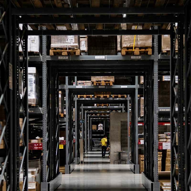 McCollisters A person in a yellow shirt walks down an aisle in a large warehouse filled with tall shelves stacked with boxes and pallets.
