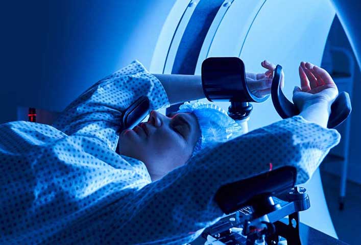 McCollisters A person wearing a hospital gown and hair cover lies on a medical imaging table with arms raised, preparing to enter an MRI or CT scanner, illuminated by blue light.