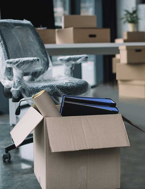 McCollisters A cardboard box filled with office supplies sits on the floor in front of a desk and an office chair wrapped in protective plastic, with several packed boxes stacked in the background, illustrating a commercial relocation.