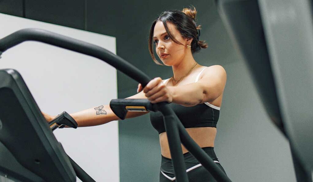 McCollisters A woman in athletic wear exercises on an elliptical machine in a gym, holding the handles and looking focused, surrounded by well-designed fitness spaces.