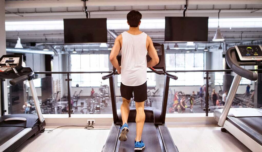 McCollisters A man wearing a white tank top and black shorts runs on a treadmill in sleek fitness spaces of a modern gym, with other equipment and people visible in the background through large windows.