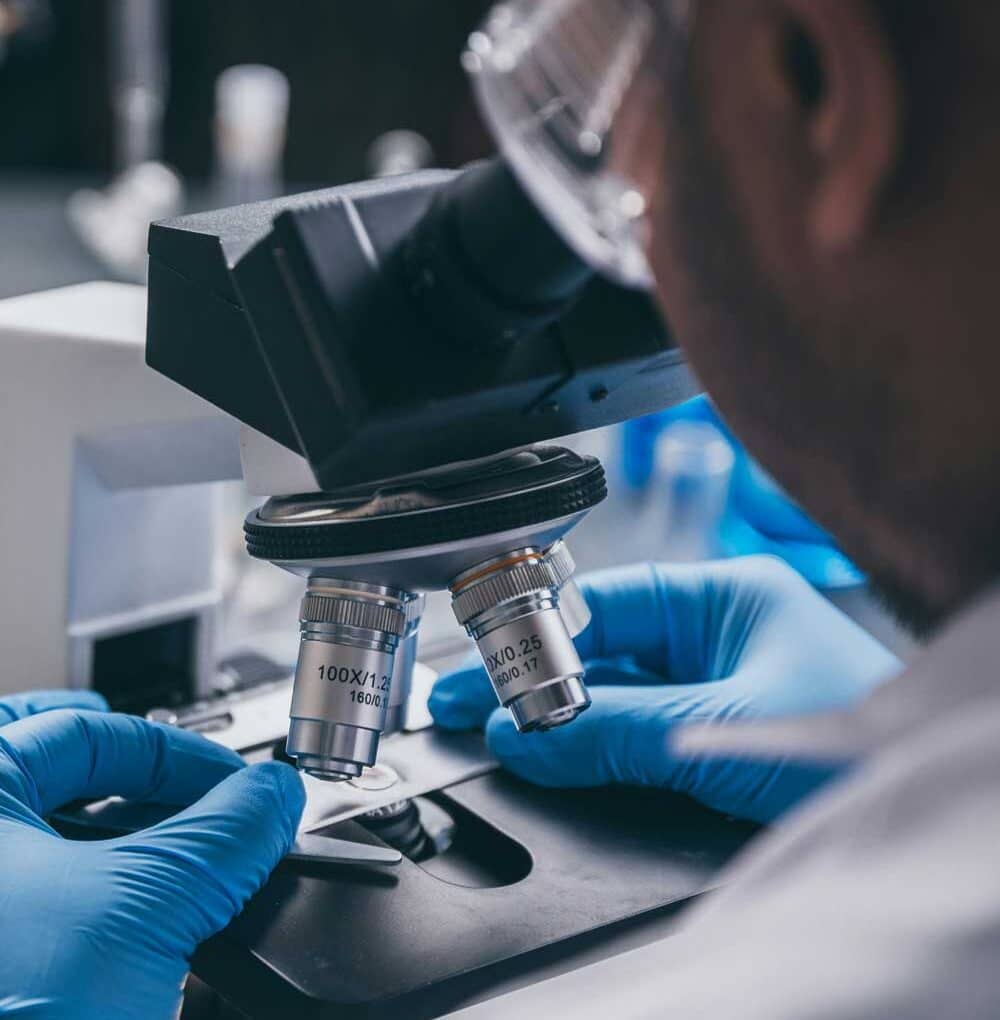 McCollisters A scientist wearing safety goggles and blue gloves adjusts a slide under a microscope, preparing for observation in a laboratory setting during a commercial relocation.