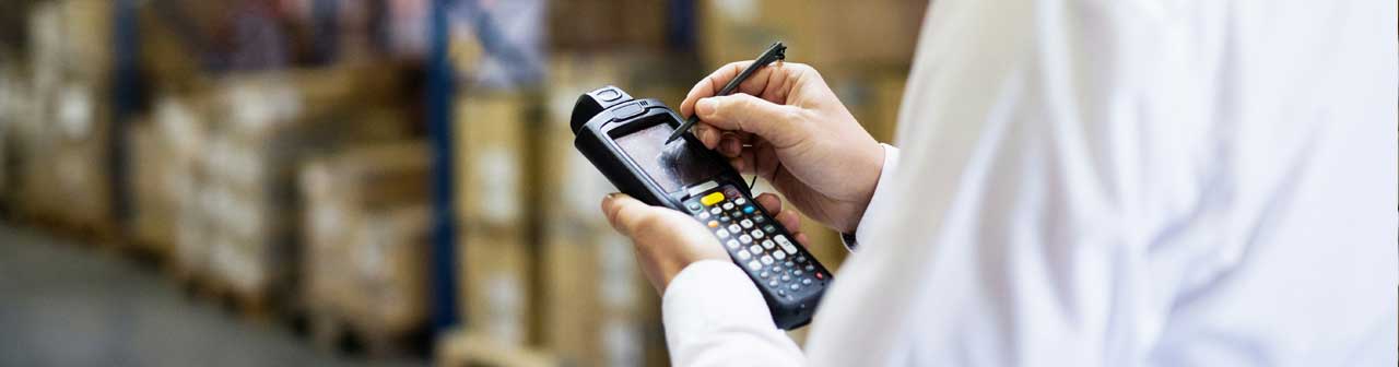 McCollisters A person in a white shirt uses a stylus to input data on a handheld barcode scanner during a commercial relocation, with shelves of boxes visible in the blurred background of a warehouse.
