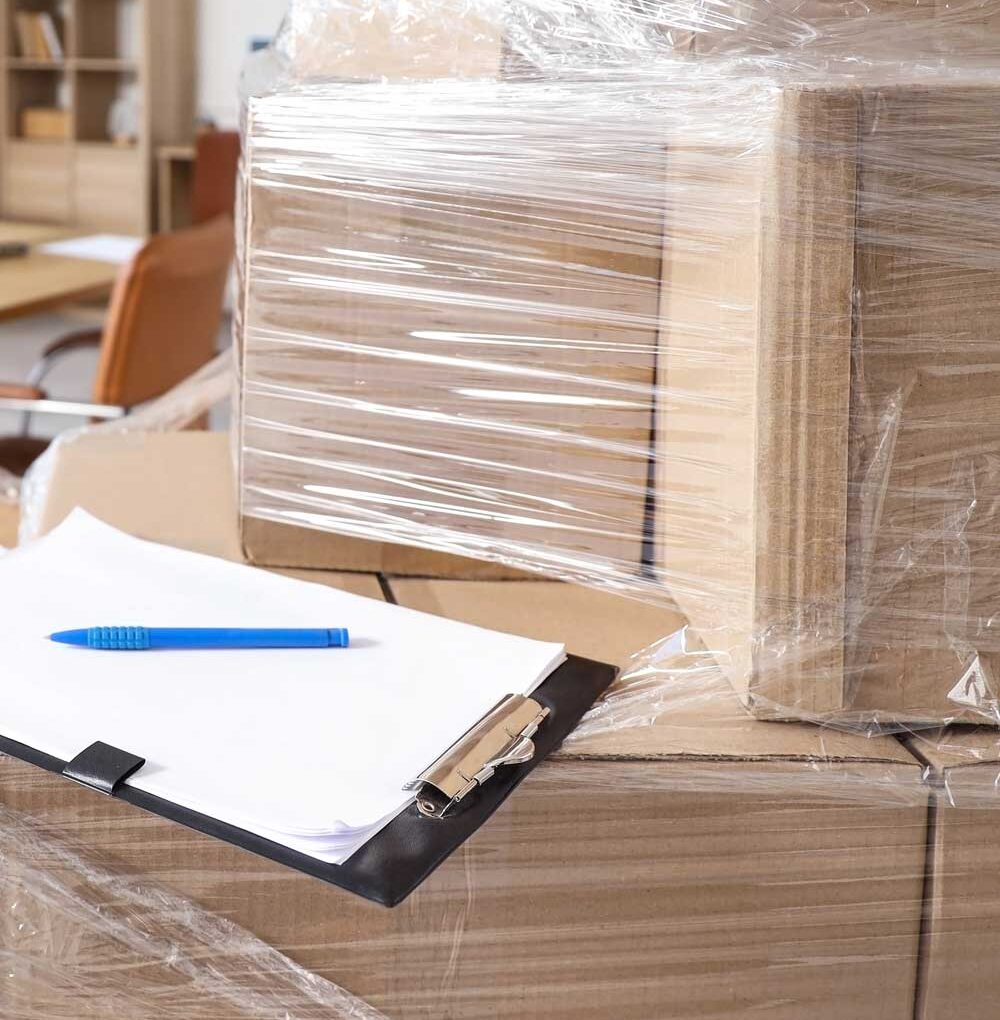 McCollisters Several cardboard boxes wrapped in plastic are stacked together, ready for commercial relocation, with a clipboard holding papers and a blue pen resting on top. The background shows an office setting with shelves and a chair.