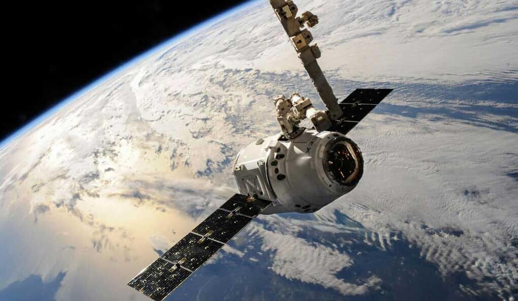 McCollisters A satellite or spacecraft, supported by an aerospace logistics partner, orbits above Earth with solar panels extended and the planet’s cloud-covered surface visible in the background.