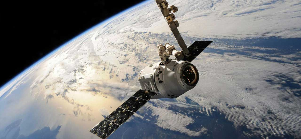 McCollisters A satellite or spacecraft, supported by an aerospace logistics partner, orbits above Earth with solar panels extended and the planet’s cloud-covered surface visible in the background.